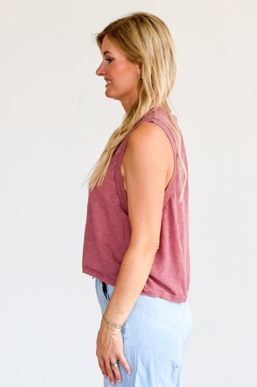 Woman wearing a pink sleeveless top and light blue pants against a white wall.