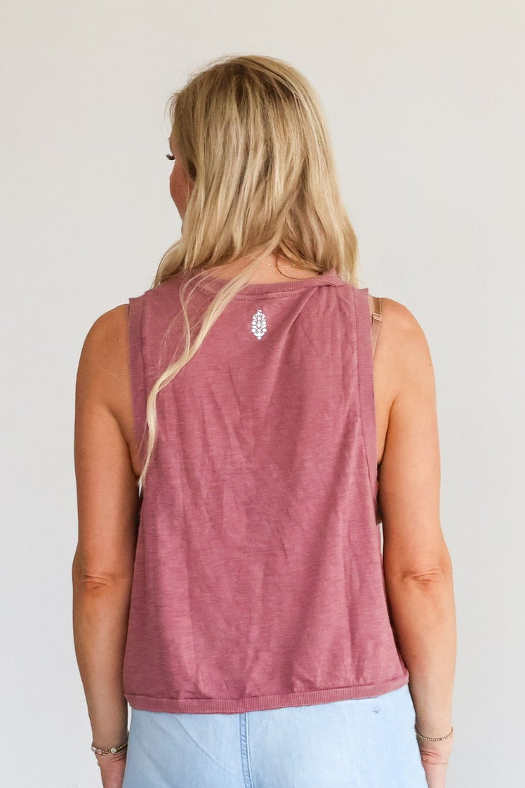 Woman wearing a pink sleeveless top and light blue pants against a white wall.